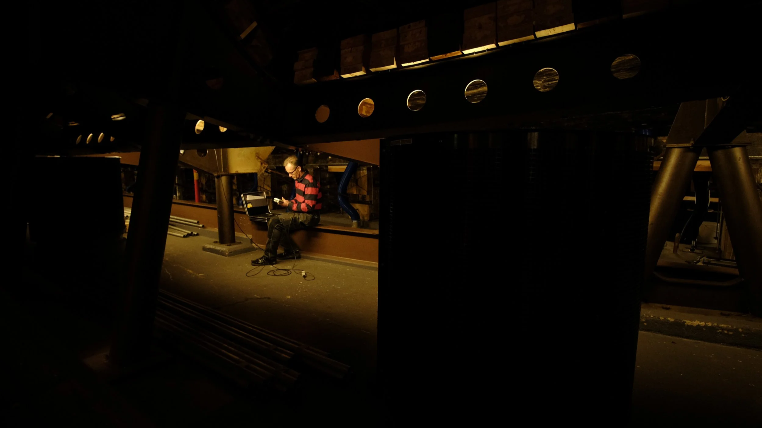 Person sitting in a dimly lit industrial space, using a laptop with cables around.