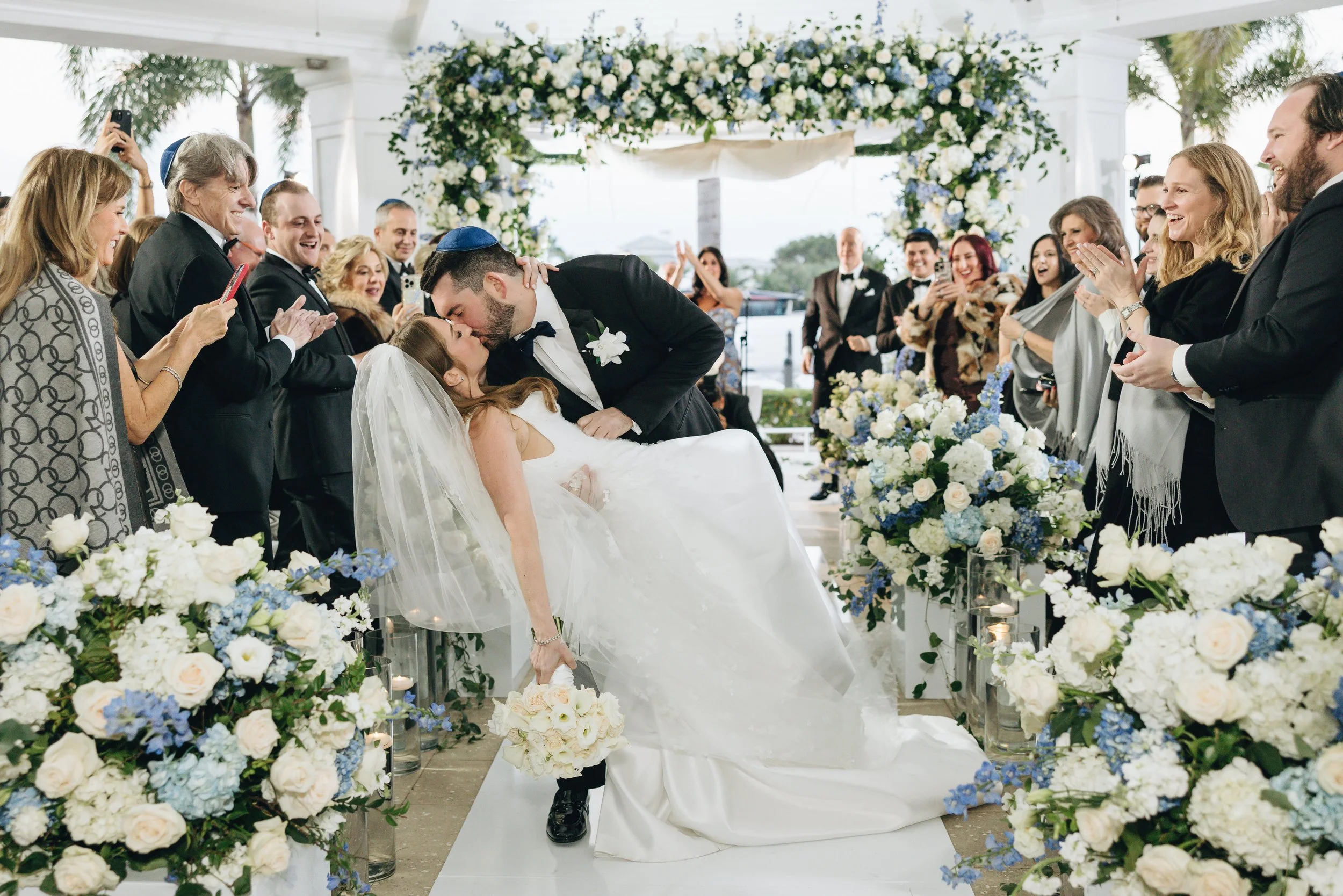 A wedding scene where a groom is kissing a bride who is leaning back, surrounded by onlookers clapping and taking photos, with floral arrangements and an arch decorated with flowers in the background.