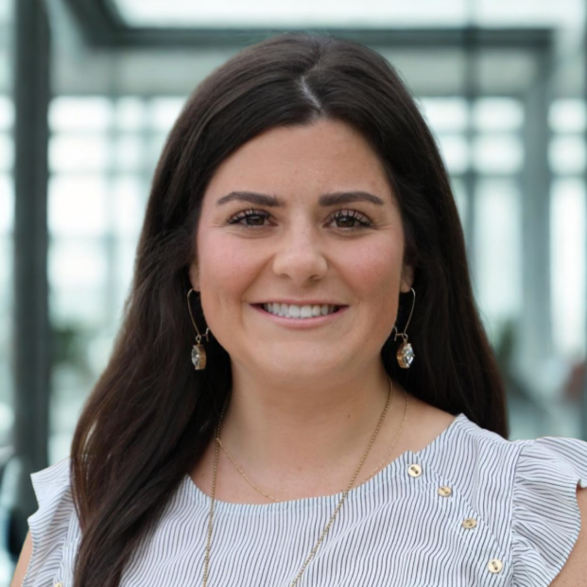 A smiling woman with dark brown hair, wearing earrings and layered necklaces, in a bright, modern indoor setting.