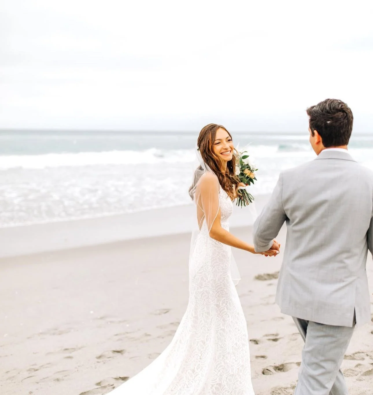 A bride in a white wedding dress and a groom in a light gray suit holding hands on the beach, smiling at each other.