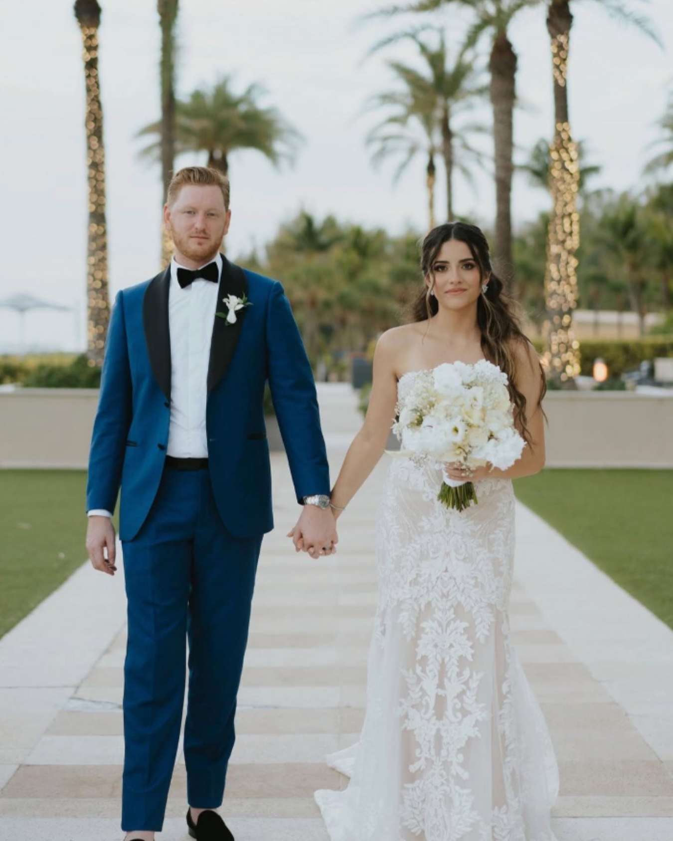 A bride and groom holding hands and walking outdoors on a wedding day, with palm trees and string lights in the background.