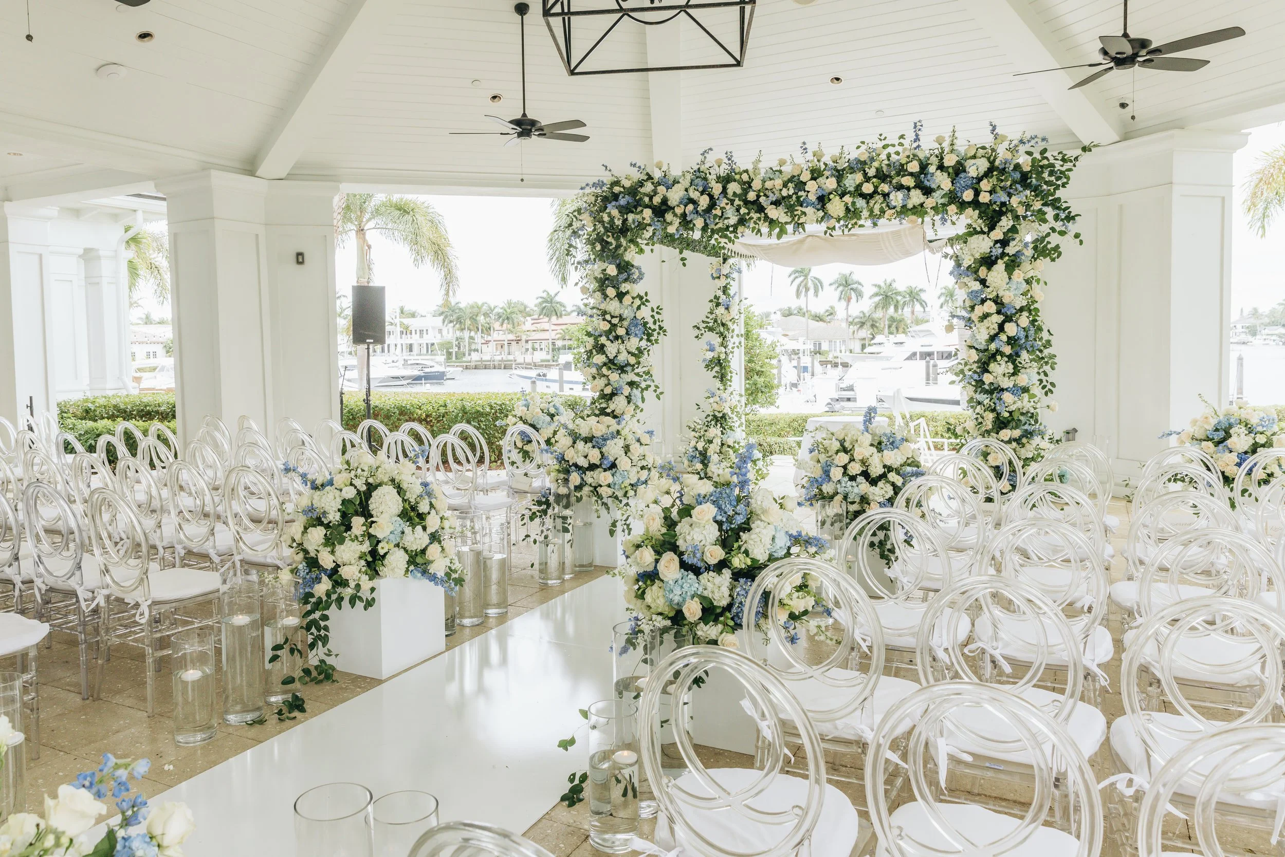 An elegant outdoor wedding ceremony setup with white floral arrangements, a floral arch, and white transparent chairs, overlooking a marina with boats and palm trees.