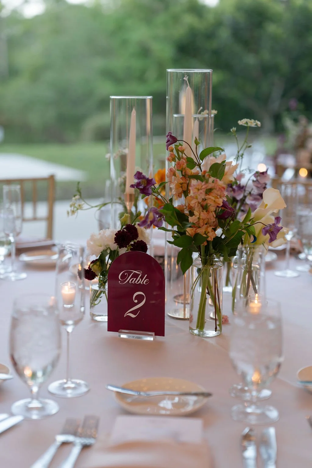 Outdoor dining table decorated with floral arrangements, candles, and candles, set up for an event, with string lights hanging overhead and trees in the background.