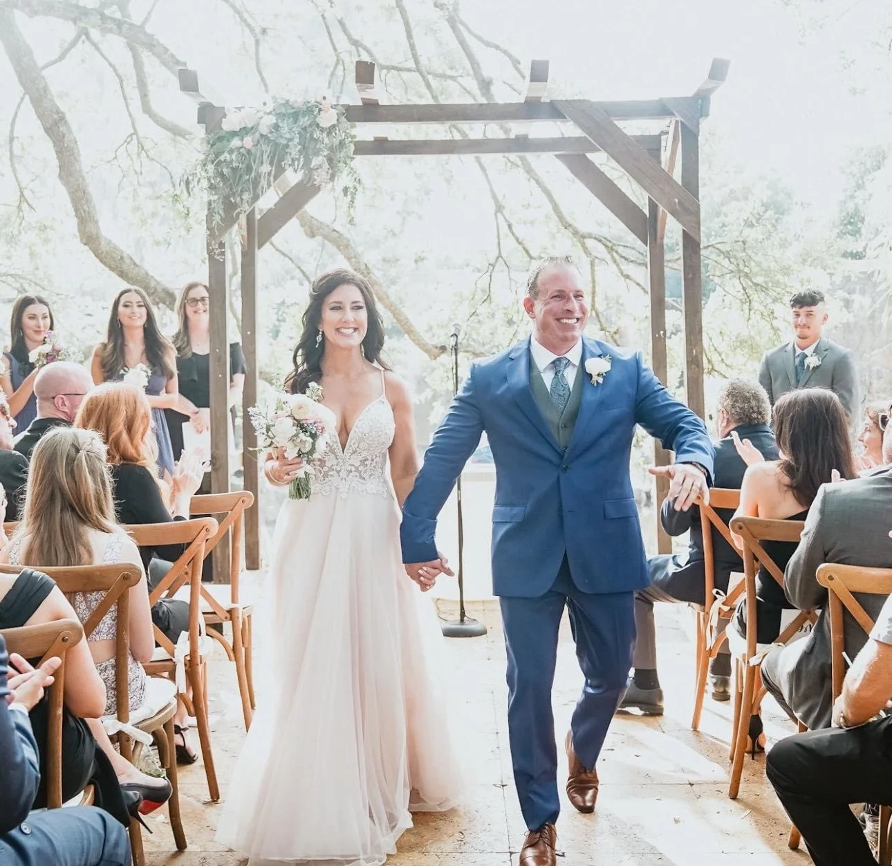 A bride and groom walking down the aisle holding hands at their outdoor wedding ceremony, surrounded by seated guests and wedding party, with a wooden arch decorated with flowers and greenery behind them.