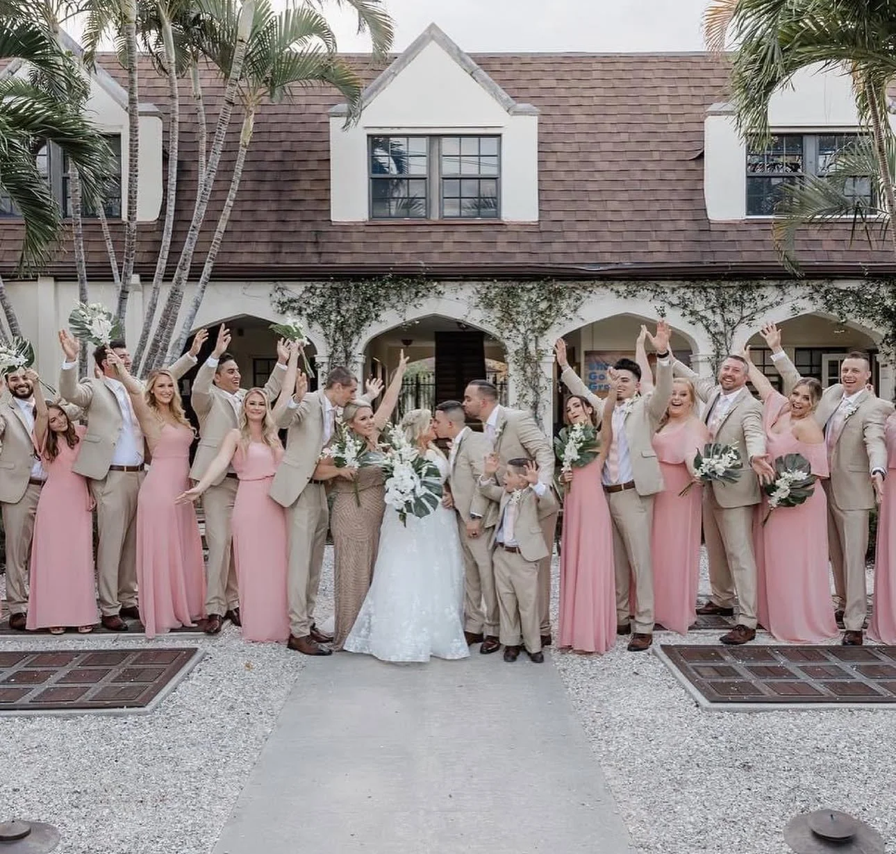 Group of bridesmaids and groomsmen celebrating outside a house, with the bride and groom kissing in the center. The bridesmaids wear pink dresses, and the groomsmen wear beige suits. They are smiling, raising their hands, and holding bouquets of flow