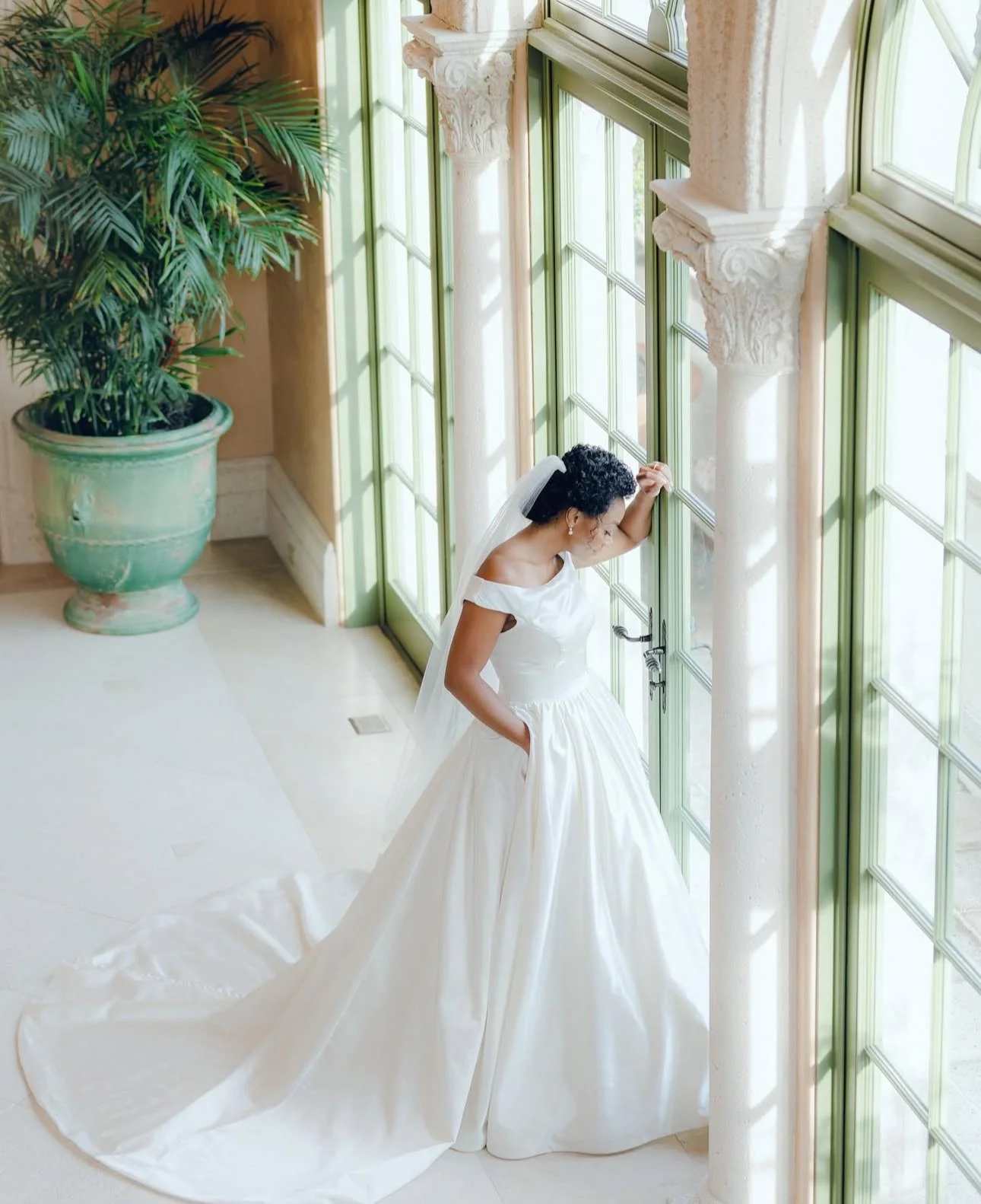 A bride in a white wedding gown standing by large green window panes, looking out with one hand on her forehead and her other hand in her pocket, with a large potted plant nearby.