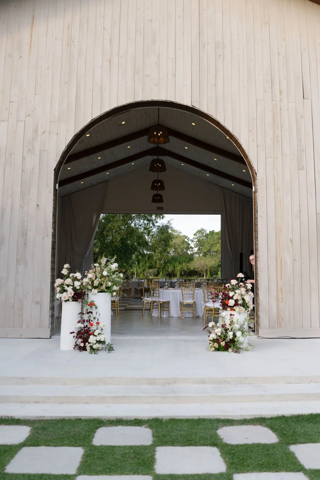 An outdoor event space with a large wooden archway and decorated with floral arrangements, leading into a reception area with round tables and gold chairs, surrounded by trees.