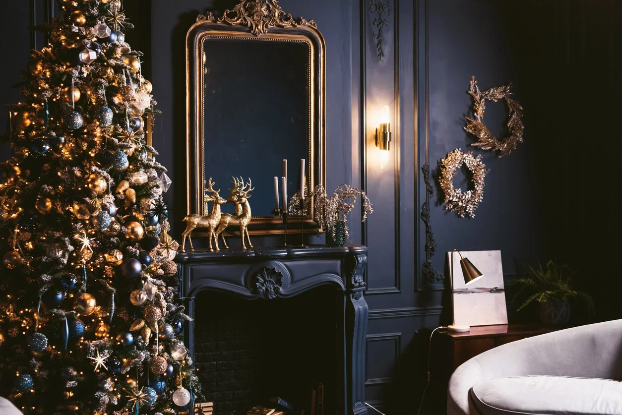 Decorated Christmas tree with gold and silver ornaments in a festive living room with dark blue walls, gold wreaths, a mirror, and holiday decorations.