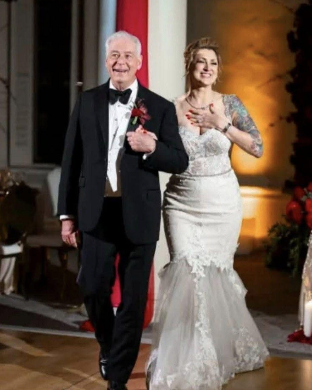 A bride and an elderly man, possibly her father, walking down the aisle at a wedding. The bride is smiling, wearing a white lace wedding dress with tattoos on her arms, and has her hand on her chest. The man is dressed in a black tuxedo with a bow tie, smiling and holding his chest with one hand.