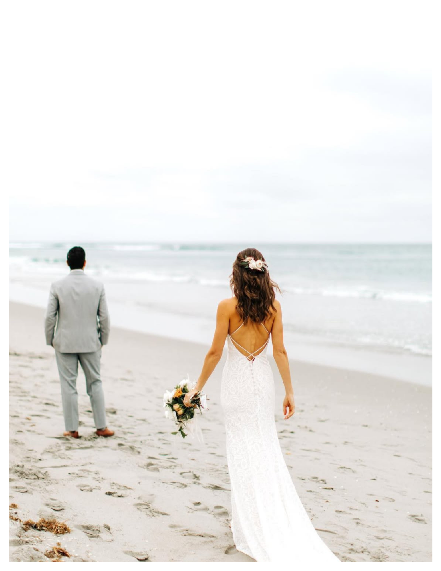 A bride and groom standing on a beach, with the bride in a white lace wedding dress holding a bouquet, and the groom in a light gray suit facing the ocean.