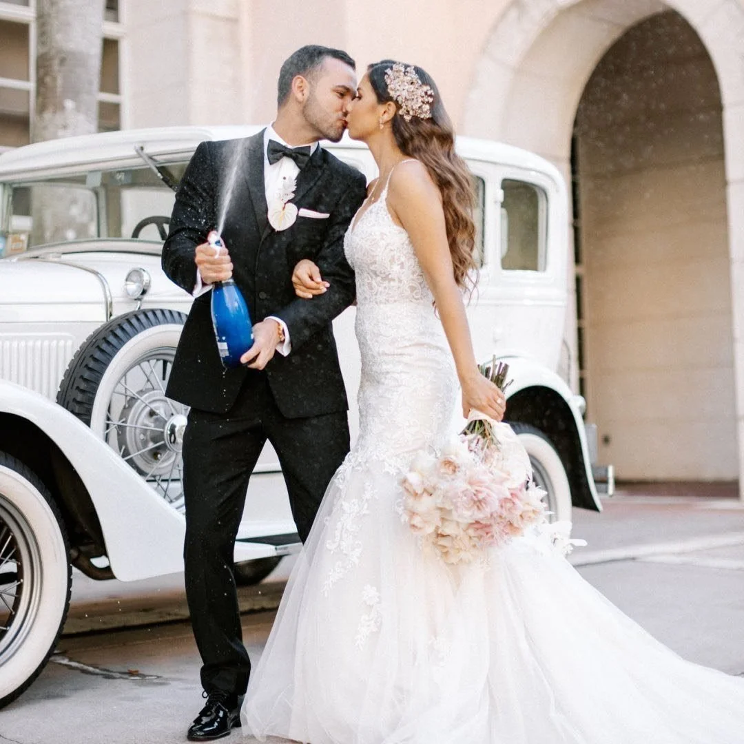 A newlywed couple kissing in front of a vintage white car. The bride is holding a bouquet and wearing a lace wedding dress with floral hair accessories, while the groom is dressed in a black tuxedo with a bow tie, holding a champagne bottle.
