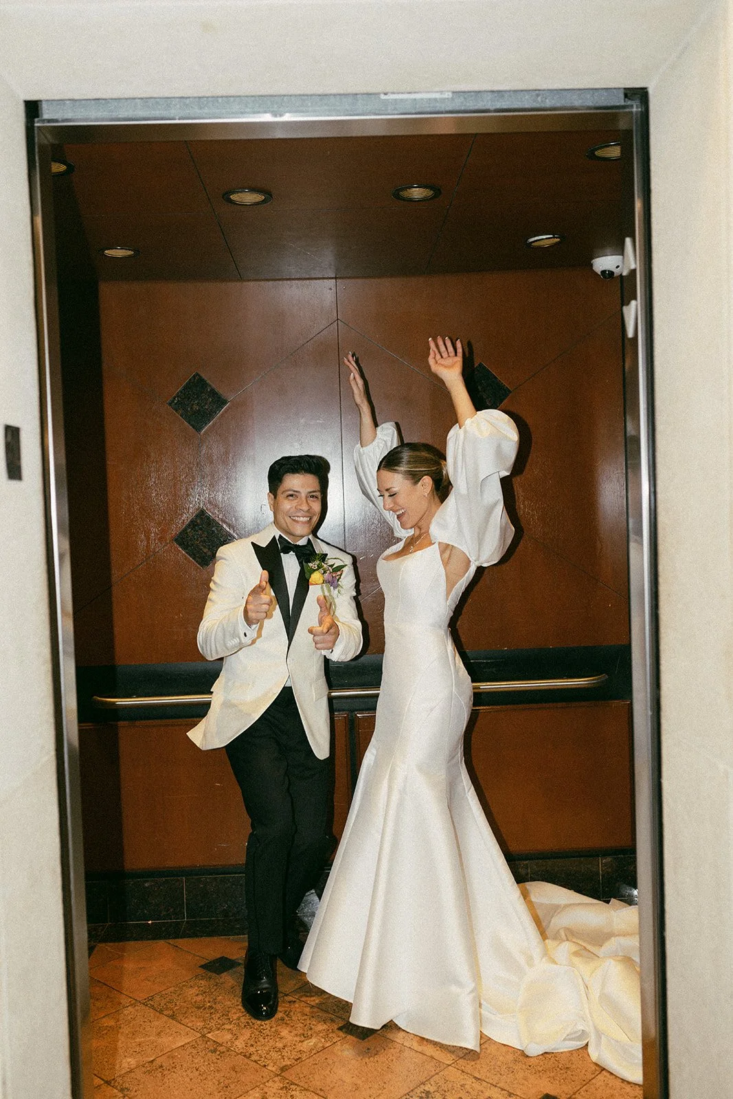 A celebrating bride and groom standing inside an elevator, with the bride raising her arms joyfully and the groom giving two thumbs up, both dressed in wedding attire.