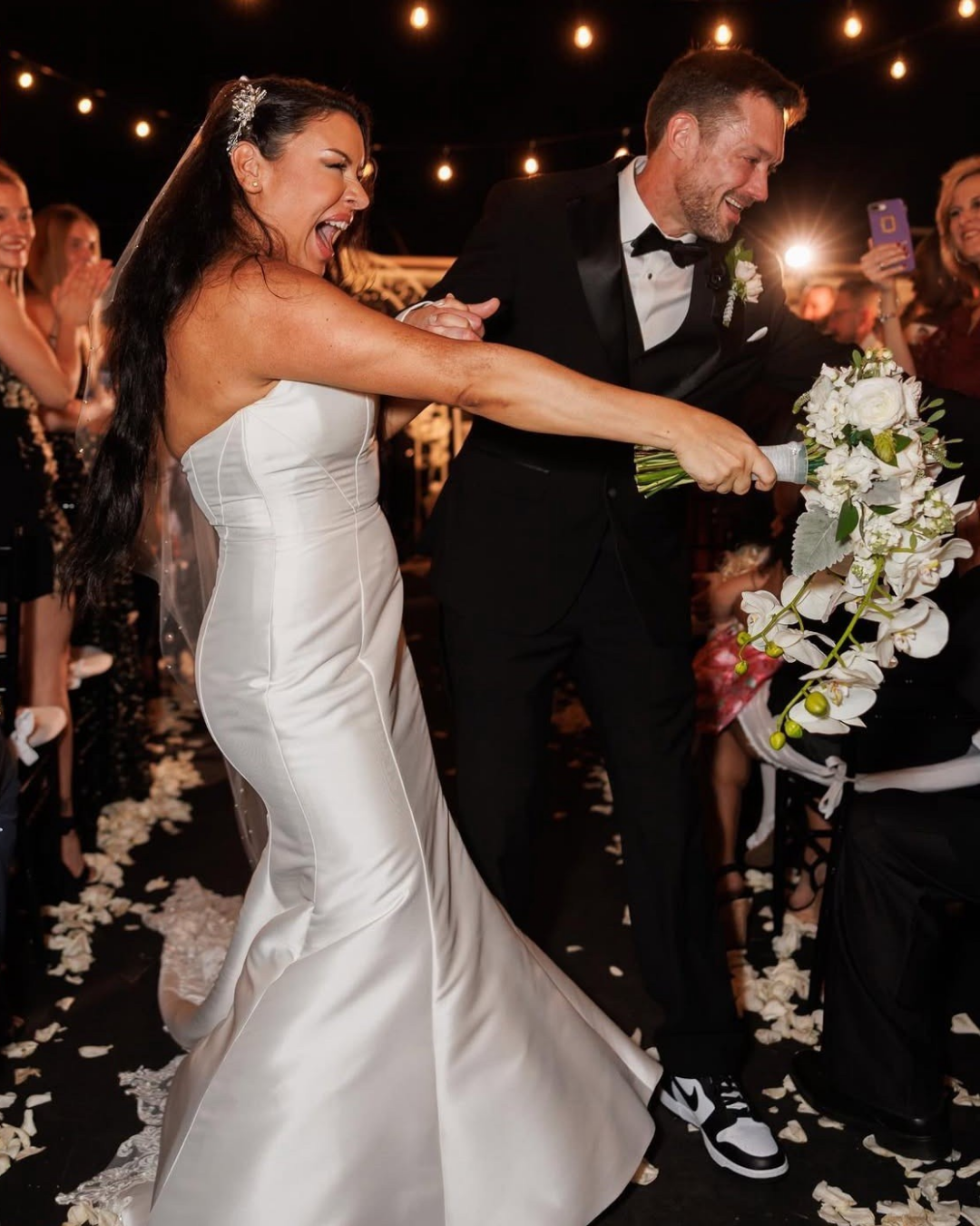 A bride and groom participate in a bouquet toss during a wedding celebration, surrounded by guests under string lights.