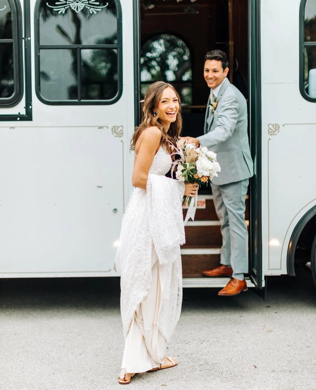 A woman in a wedding dress holding a bouquet, smiling, standing outside near a white vintage bus, with a man in a light gray suit standing in the doorway of the bus, also smiling.