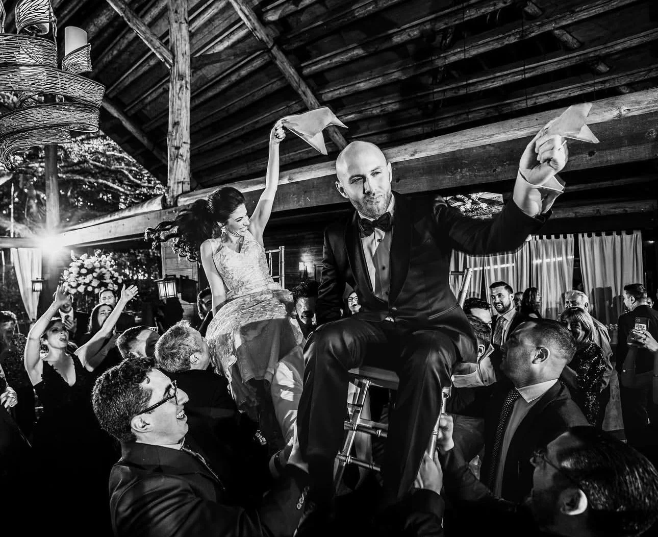 Black and white photo of a lively wedding reception with a man in a tuxedo sitting on a chair, holding a napkin, surrounded by guests celebrating, some seated and others dancing, in a rustic venue with wooden beams.