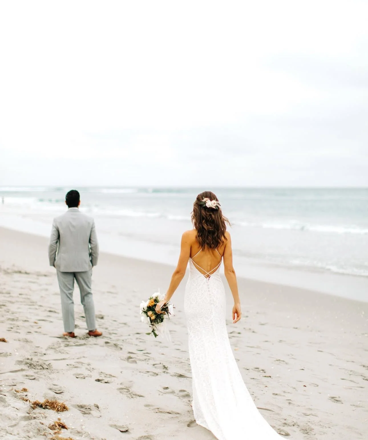 Bride in a white lace wedding dress holding a bouquet walking on the beach with a groom in a gray suit at a distance.