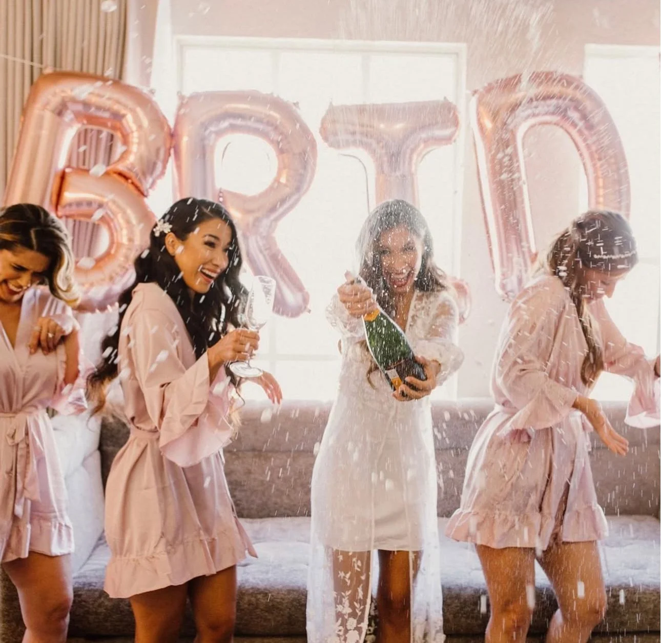 Four women celebrating a bachelorette party, wearing matching robes, with balloons spelling 'BRIDE' in the background, and champagne popping among them.