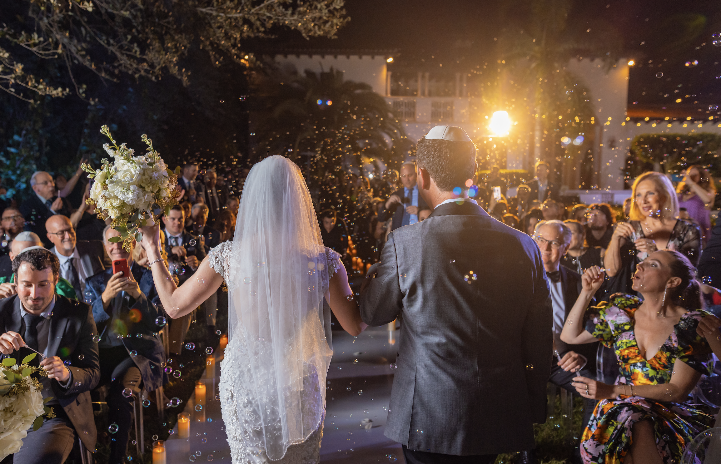 A bride and groom walking down the aisle at their wedding ceremony, surrounded by guests, with the bride holding a bouquet and wearing a white lace wedding dress and veil, during the evening outdoor celebration illuminated by warm lighting.