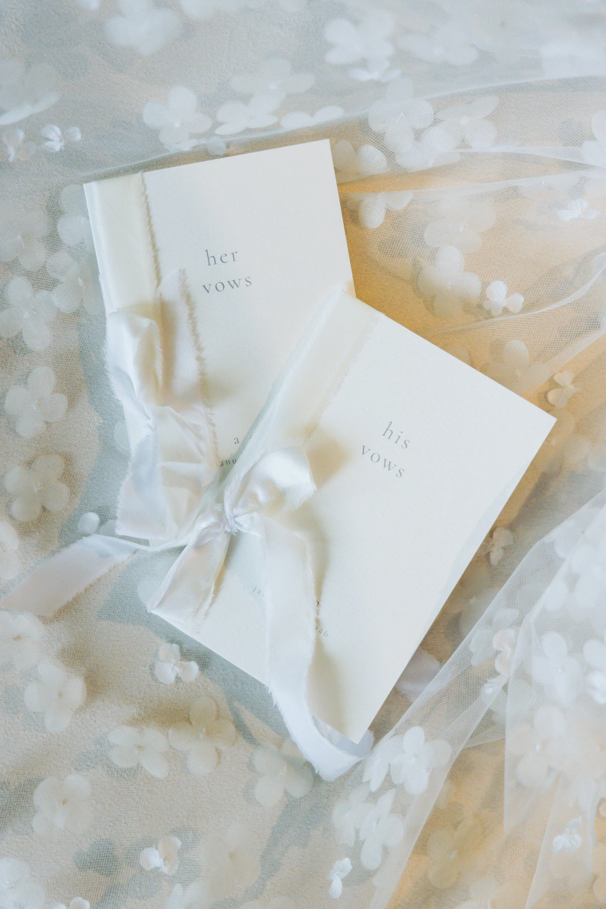 Two white vow booklets with torn edges, one labeled 'her vows' and the other 'his vows,' tied together with a white satin ribbon, surrounded by sheer fabric with small white floral patterns.