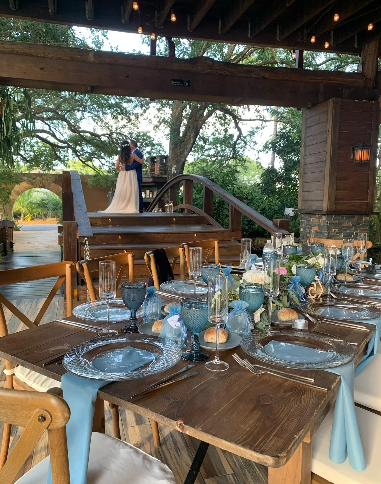 Decorated outdoor dining table set for a wedding reception with blue and white tableware, flowers, and bread, with a bride and groom dancing on a wooden stage in the background under string lights.