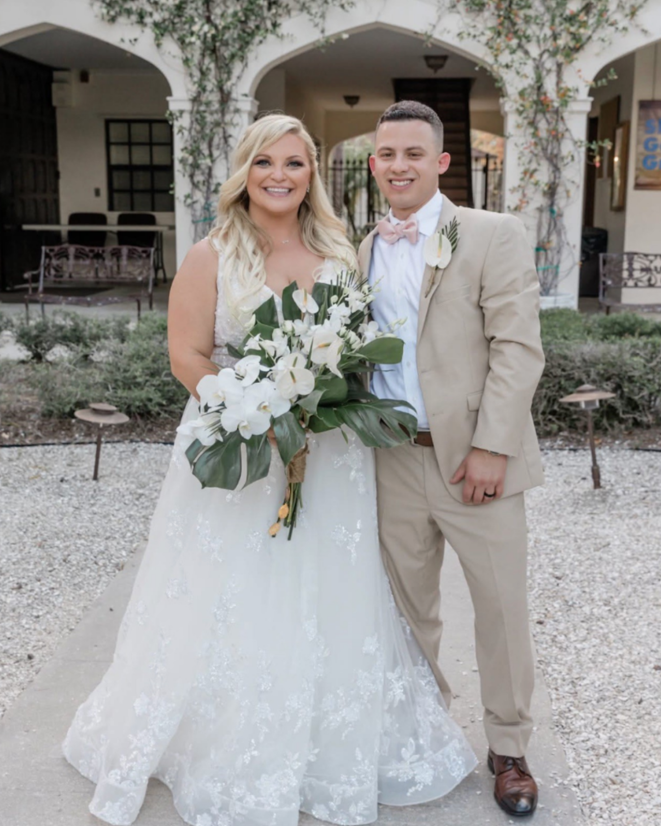 A bride and groom standing outside a building, smiling at the camera. The bride is holding a bouquet of white flowers with green leaves, and the groom is wearing a beige suit with a pink bow tie and a white shirt.