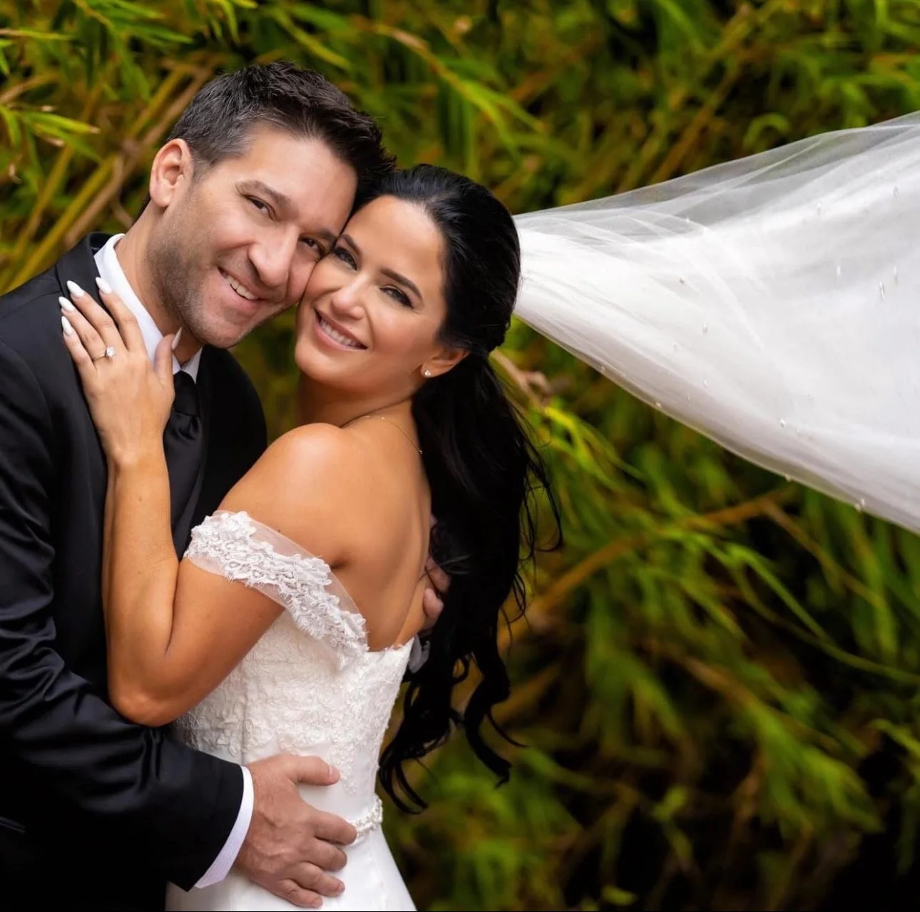 A smiling bride and groom embracing outdoors with green foliage in the background. The bride's veil blows in the wind, and she wears an off-the-shoulder white wedding dress. The groom wears a black tuxedo.