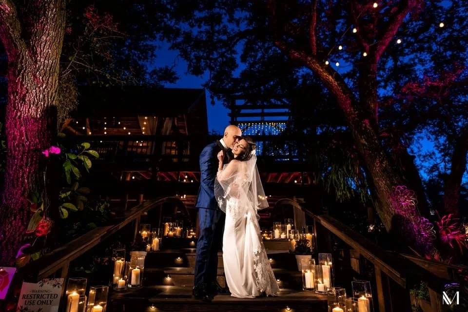 A bride and groom sharing a kiss on an outdoor staircase decorated with candles and string lights, trees with pink and purple lights surrounding them at night.