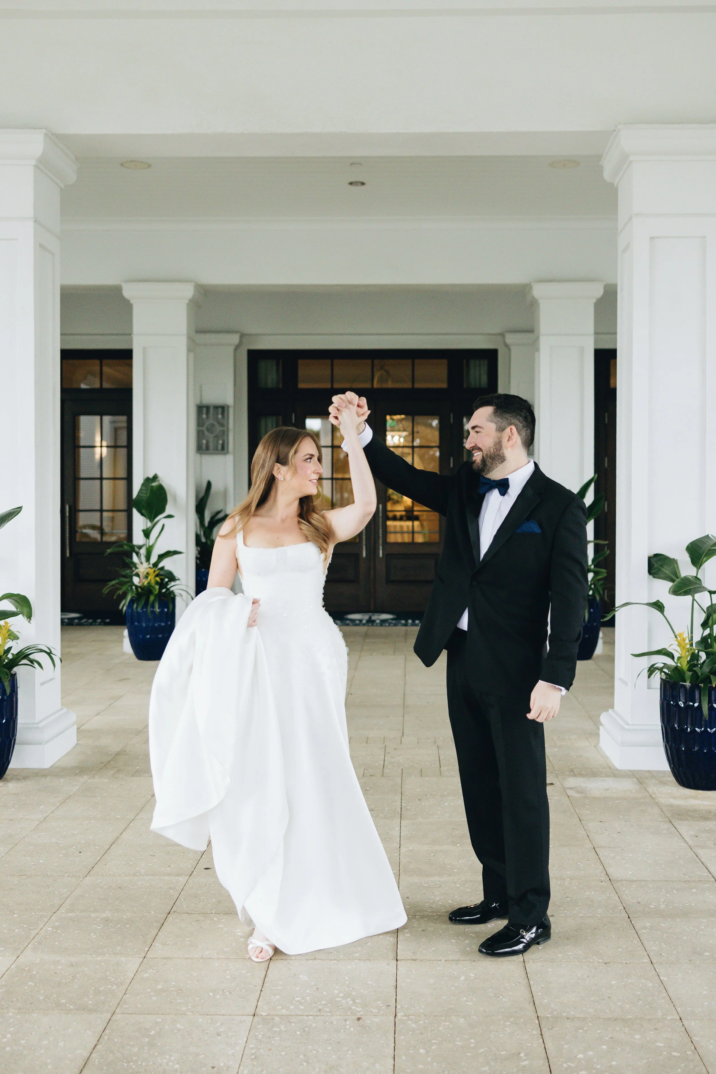 A bride and groom dancing inside a bright, elegant lobby with white columns and large windows, holding hands and smiling at each other.