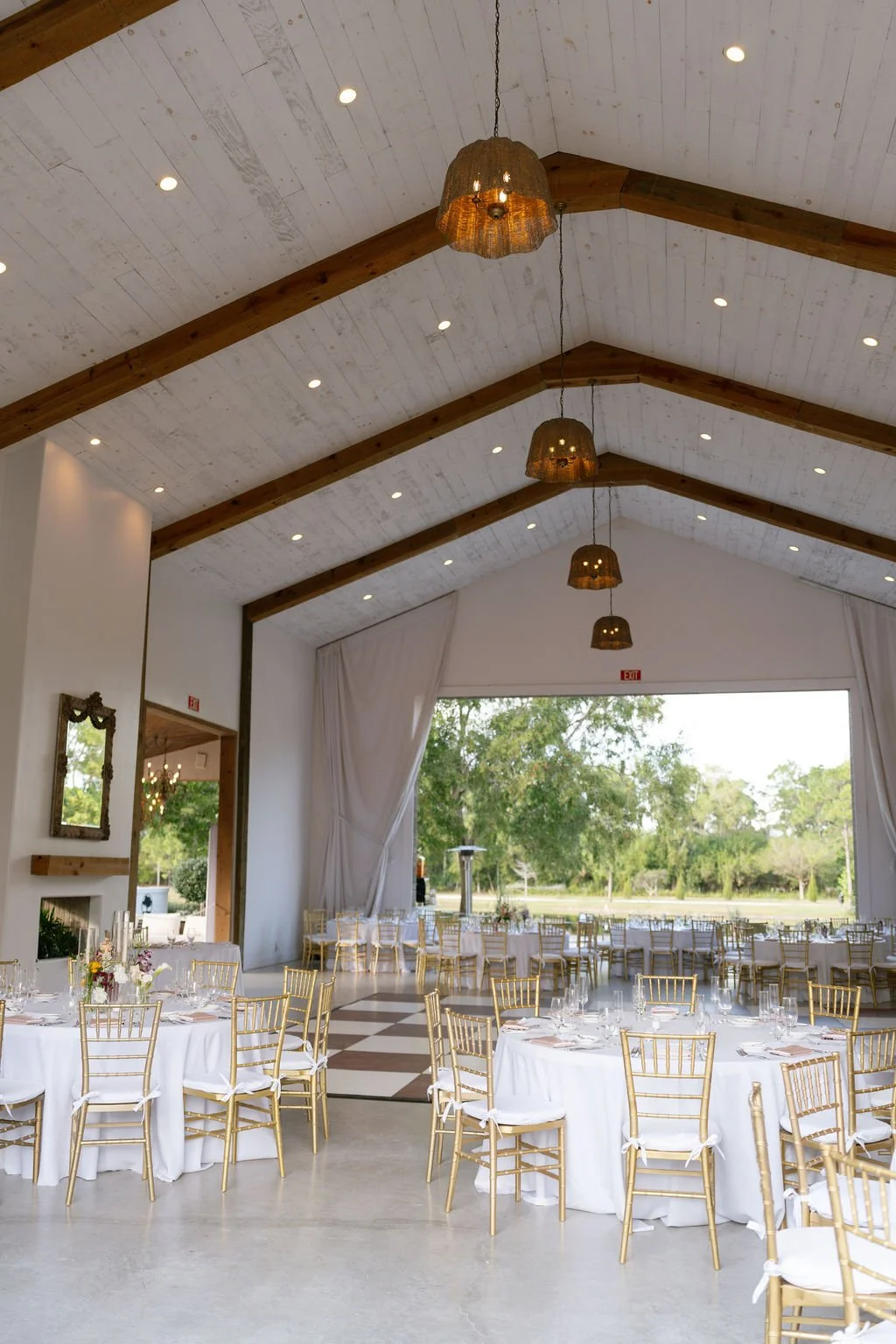 An elegant event space with round tables dressed in white tablecloths, gold chairs, and floral centerpieces. The ceiling features wooden beams and hanging pendant lights. Large windows reveal a scenic outdoor view with trees.