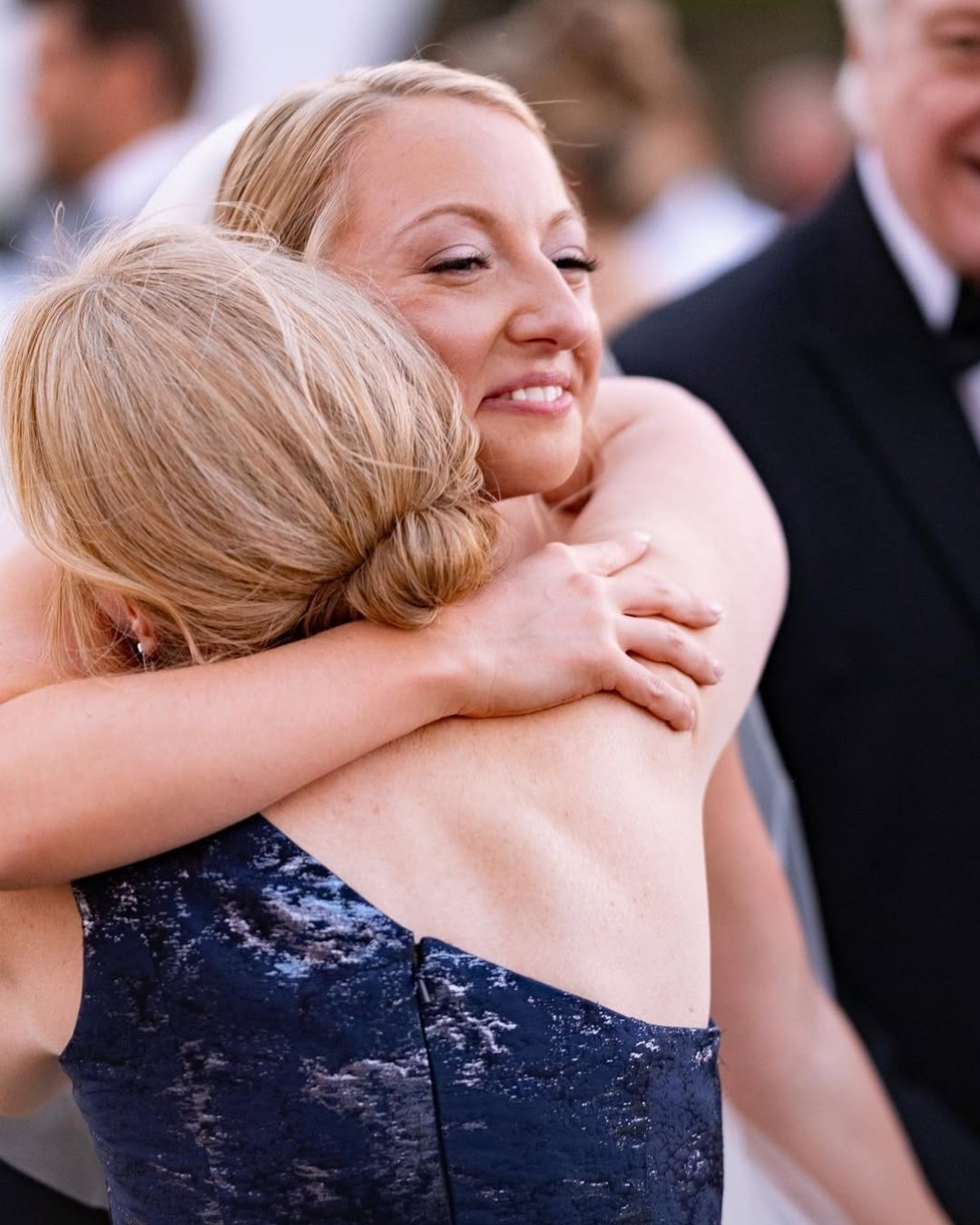 A woman with blonde hair smiling and hugging a person with red hair at a formal event, with other guests in the background.
