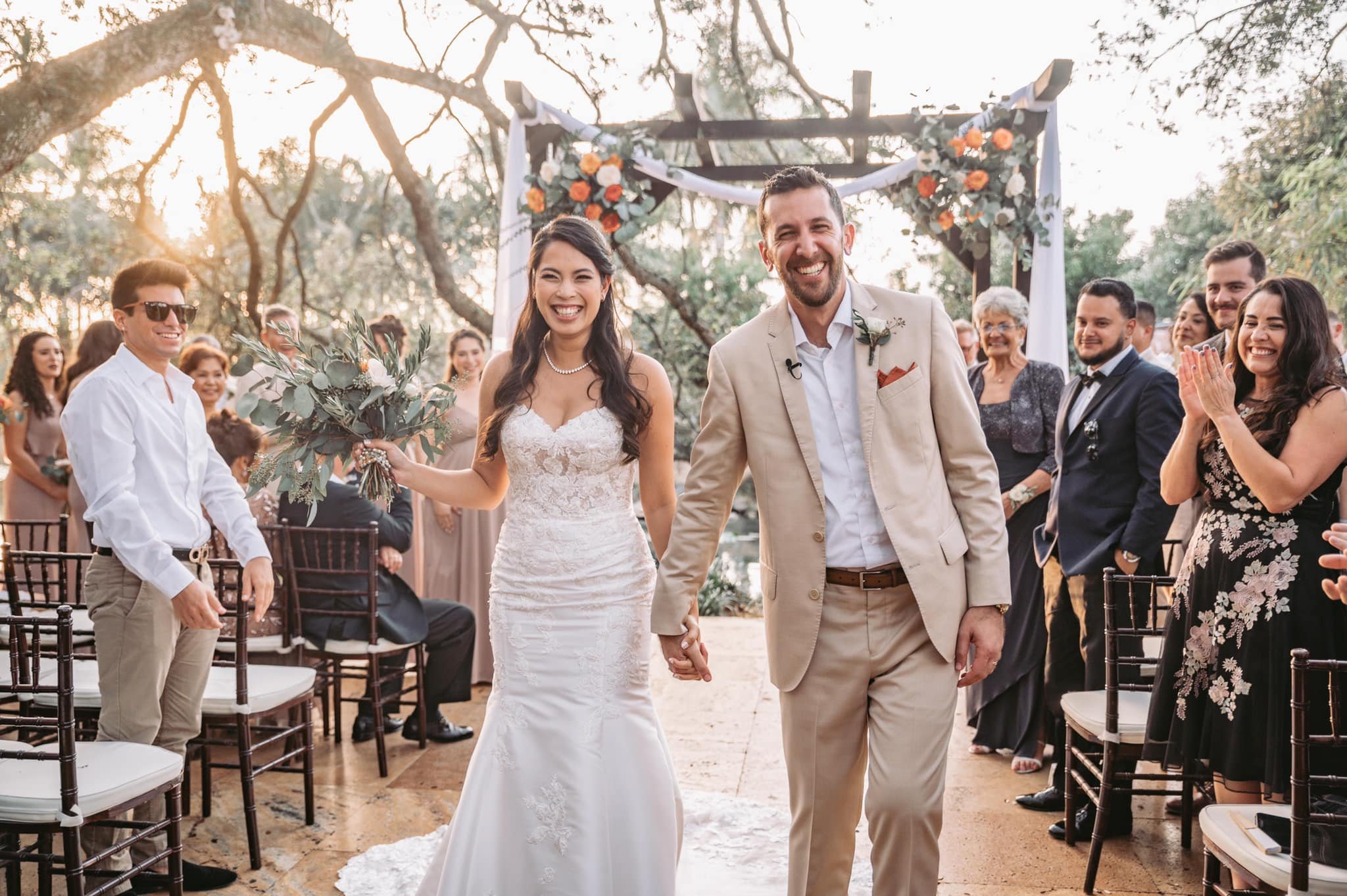 A newlywed couple walking hand-in-hand down the aisle at a wedding ceremony outdoors, surrounded by friends and family, with a floral arch behind them, during sunset.