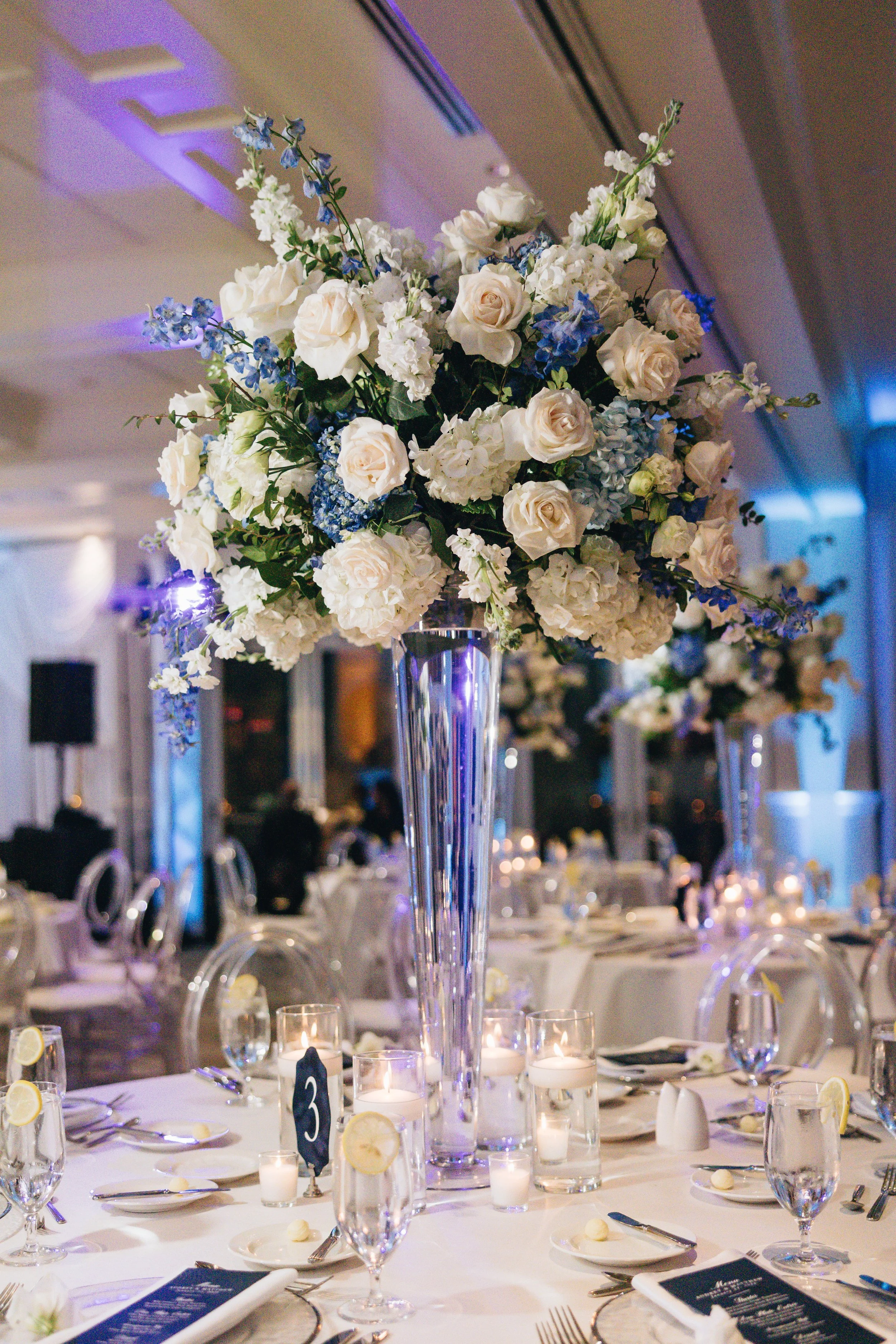 A large floral centerpiece with white roses, hydrangeas, and blue flowers in a tall clear vase on a decorated banquet table at a wedding or formal event.