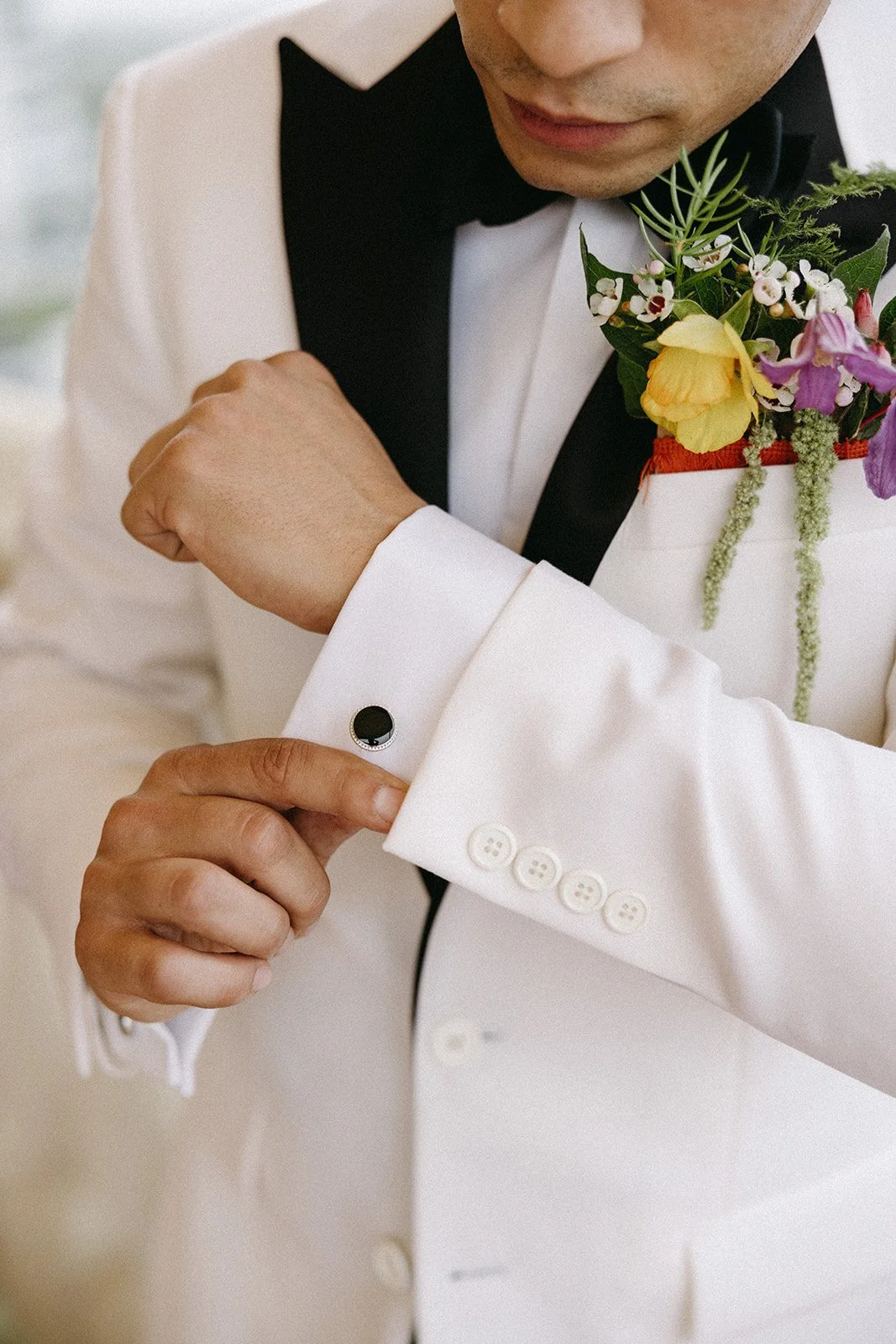 A man in a white tuxedo adjusting a cufflink on his sleeve, with a boutonnière featuring colorful flowers on his lapel.