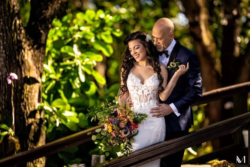 A bride and groom are standing closely together outdoors, smiling. The bride is wearing a white lace wedding dress and holding a colorful bouquet. The groom is in a dark suit with a bow tie, looking at the bride. There are lush green trees and sunlig