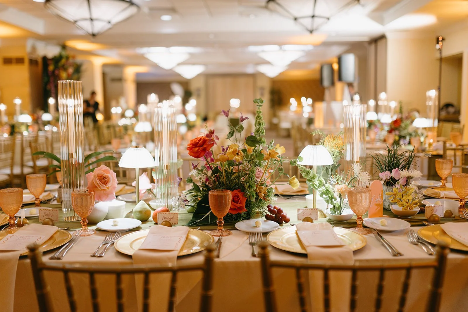 A banquet table decorated for a wedding or event with floral centerpieces, candles, place settings, and glassware in a well-lit banquet hall.