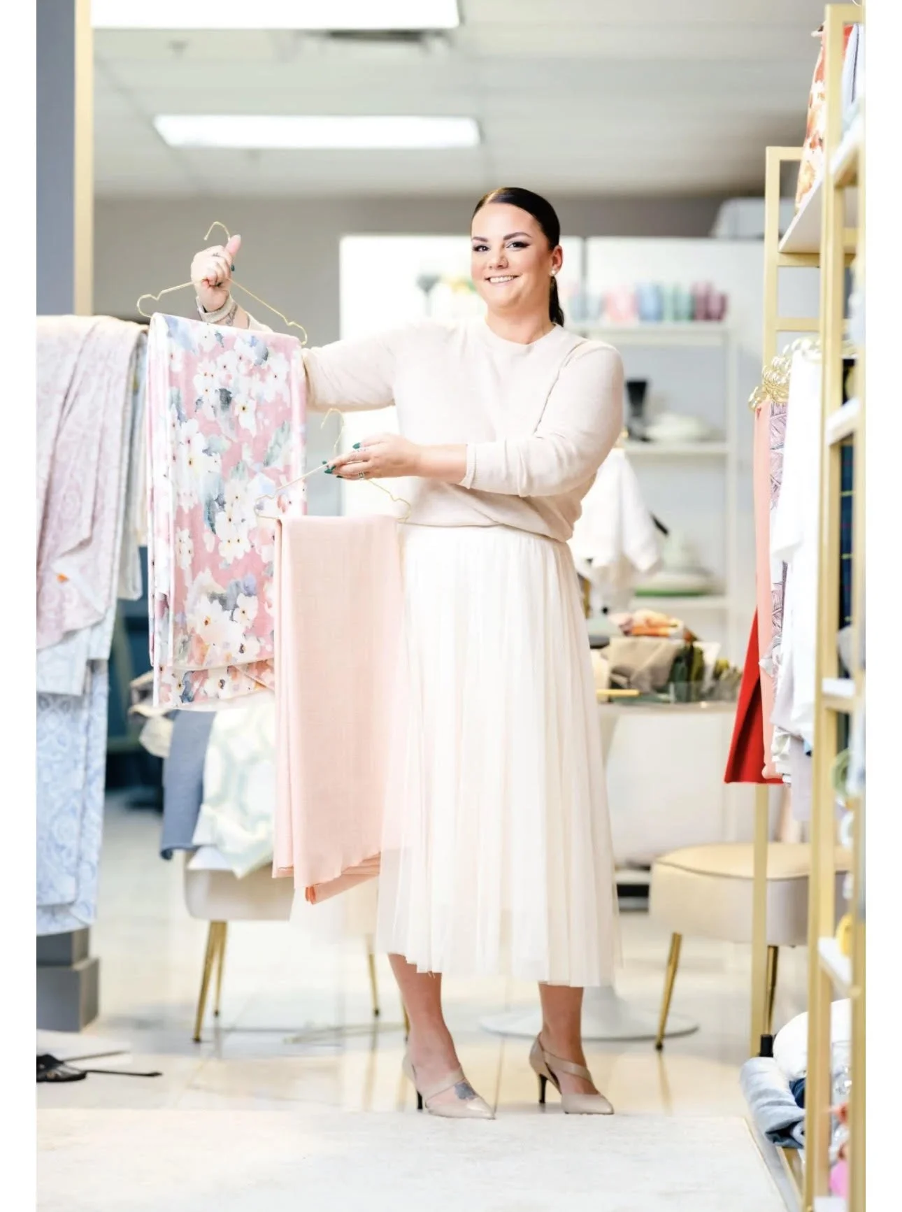 A woman with dark hair tied back, smiling, wearing a cream long-sleeve top and a cream pleated midi skirt, stands in a fabric or clothing store, holding a hanger with a pink floral garment and a fabric swatch.