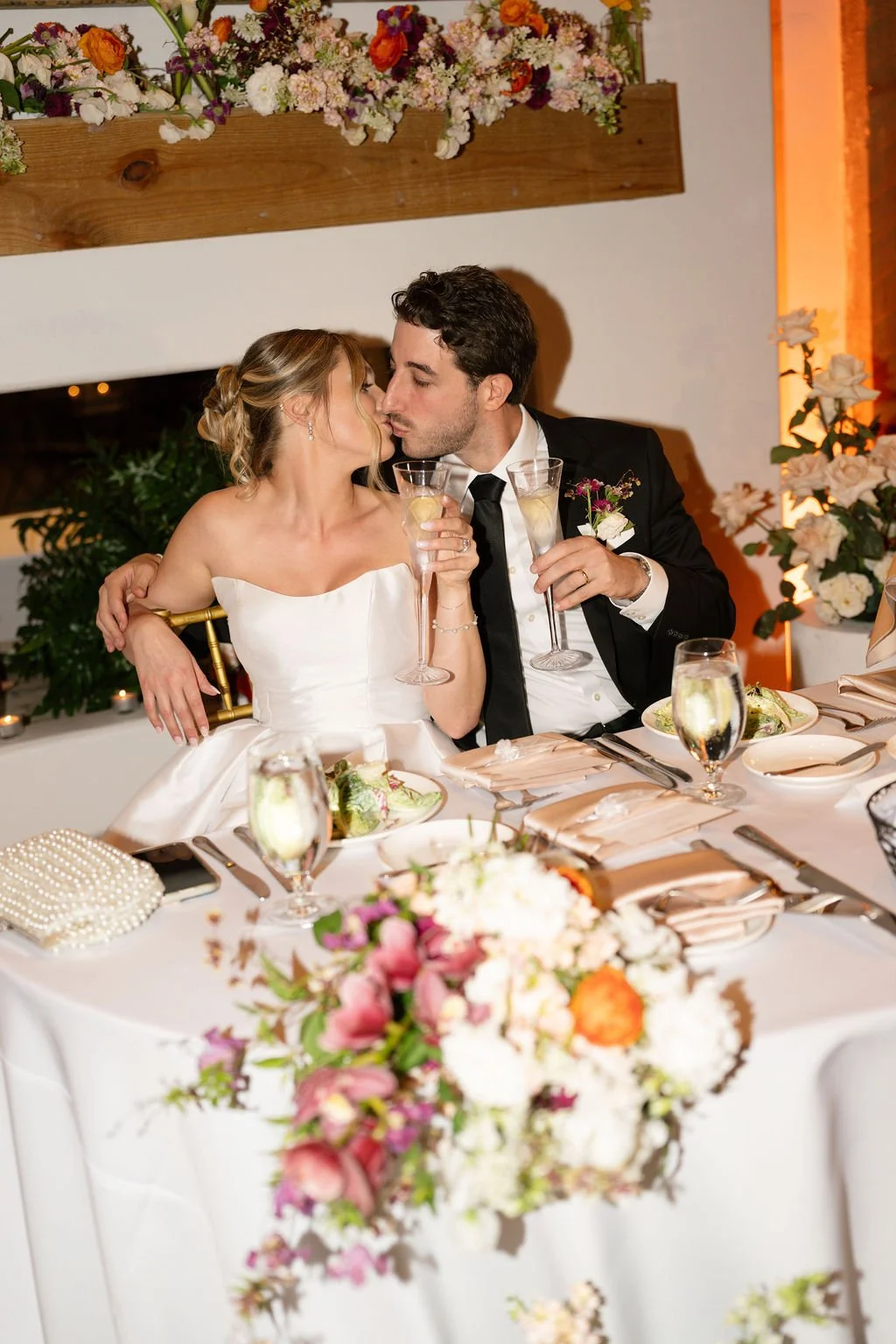 A bride with blonde hair and a floral crown smiling with a groom in a dark suit, outdoors surrounded by greenery and flowers.