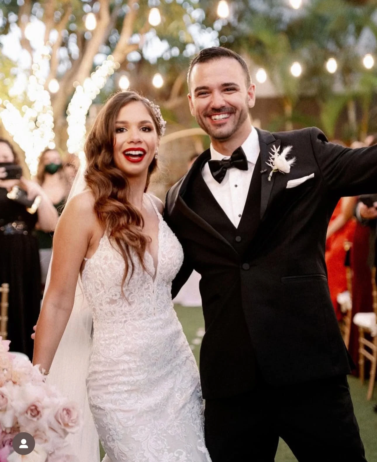 A bride and groom are smiling and taking a selfie at their wedding reception, decorated with string lights and trees.