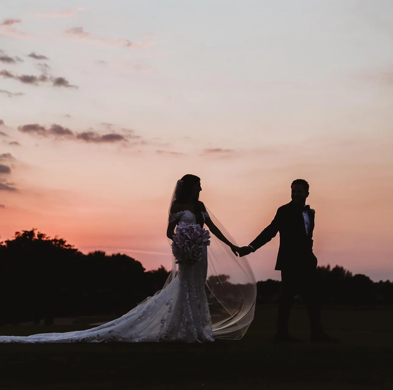 Silhouette of a bride and groom holding hands at sunset with colorful sky.