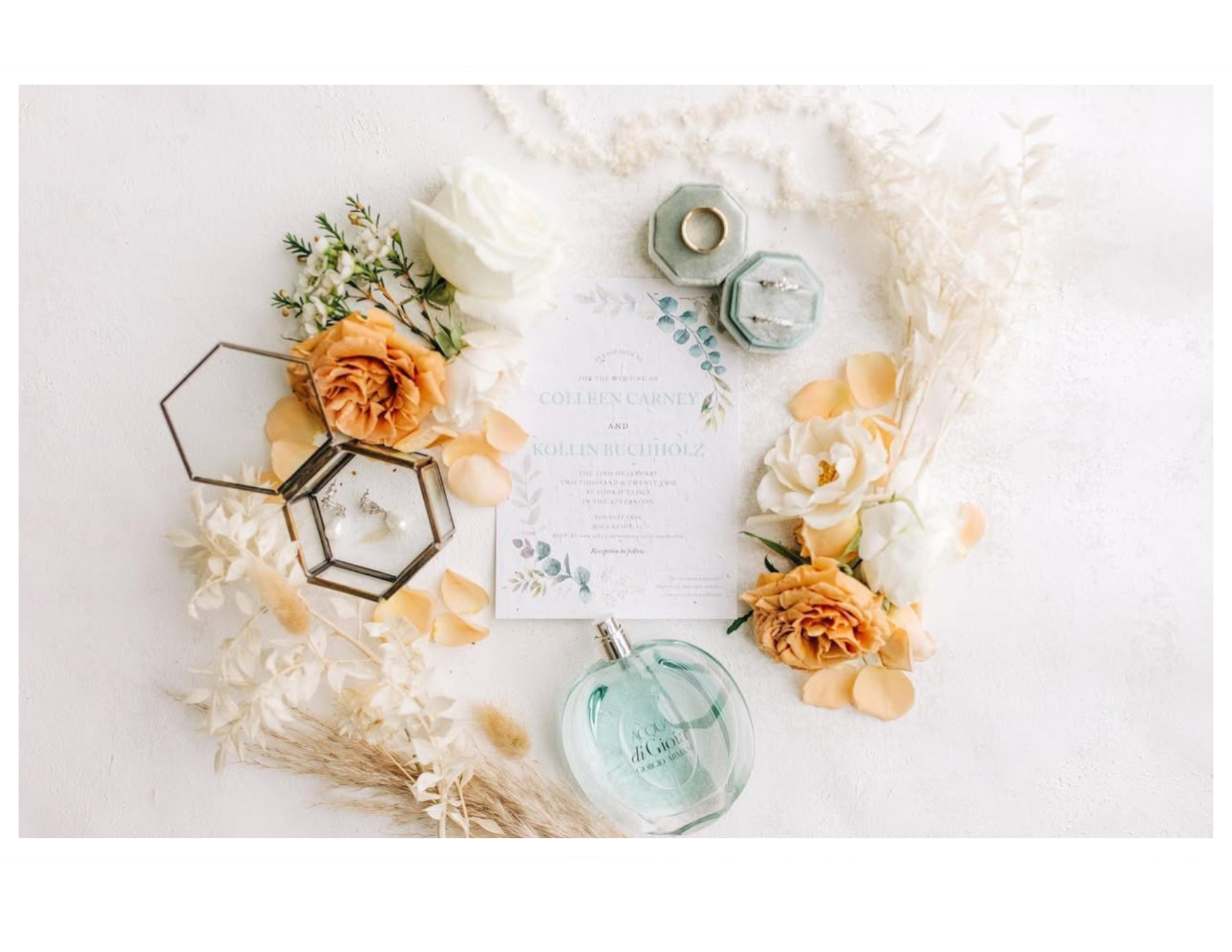 Wedding invitation surrounded by flowers, rings, perfume, and decorative jars on a white surface.