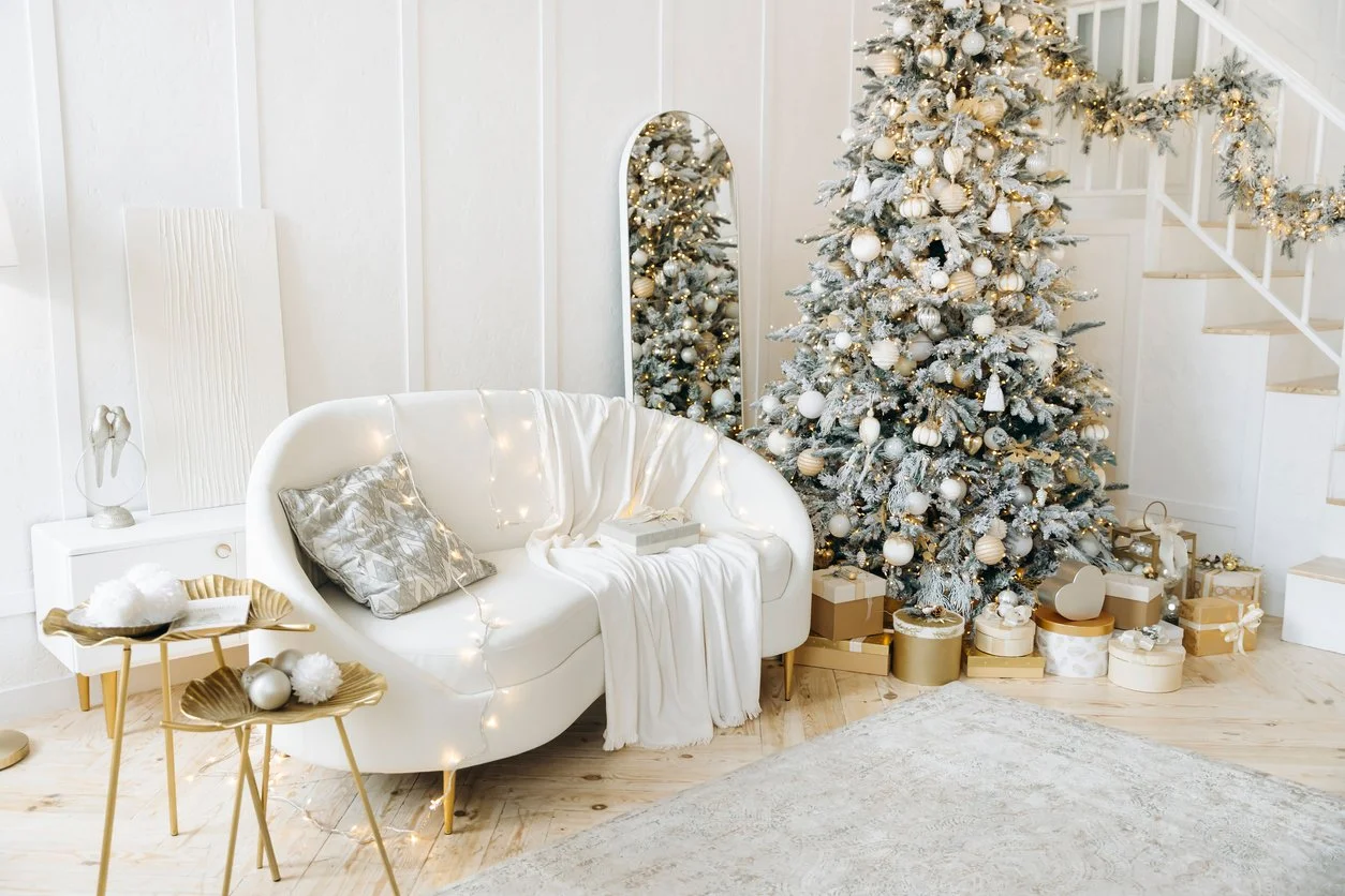 A decorated Christmas tree with white ornaments and gold accents, surrounded by wrapped presents. A white sofa with a cushion, blanket, and string lights is nearby, along with two gold side tables holding decorative items. The room has white walls and hardwood floors, creating a festive holiday scene.
