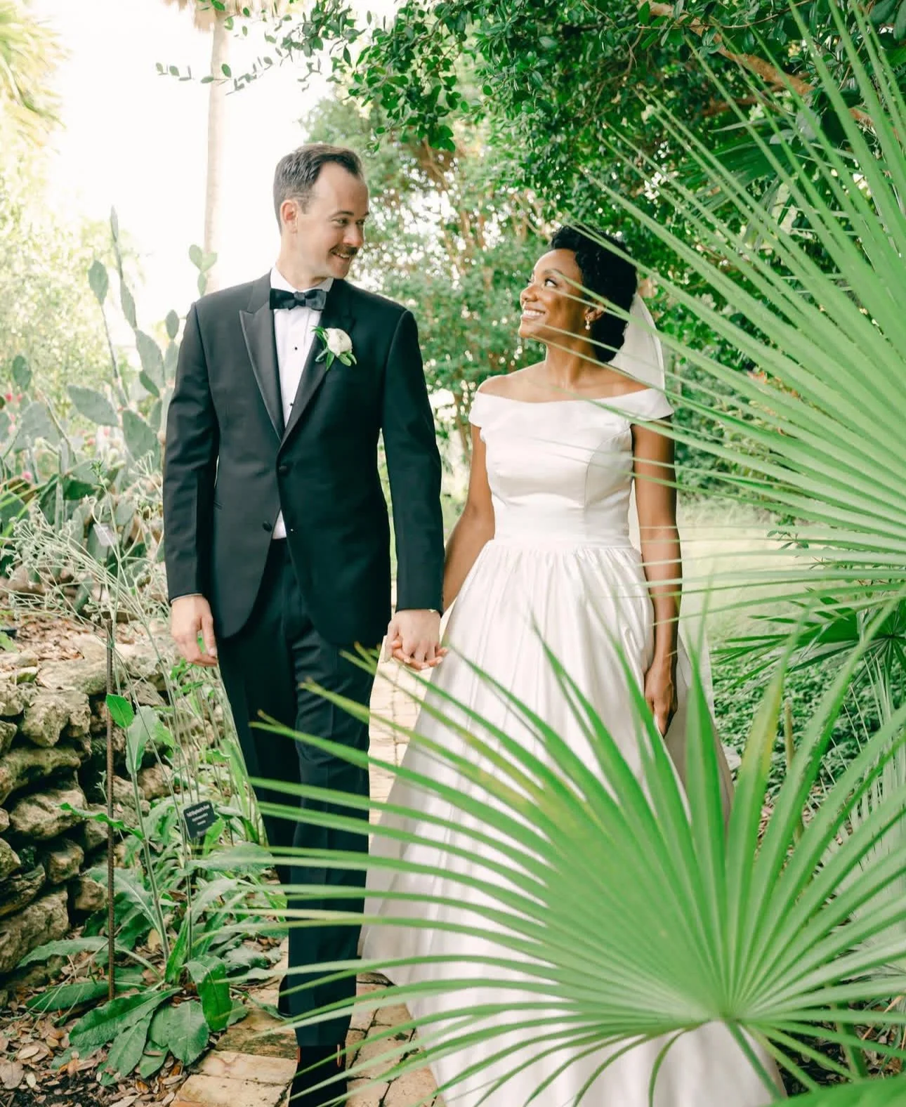 Bride and groom holding hands in a lush green garden setting, smiling at each other.