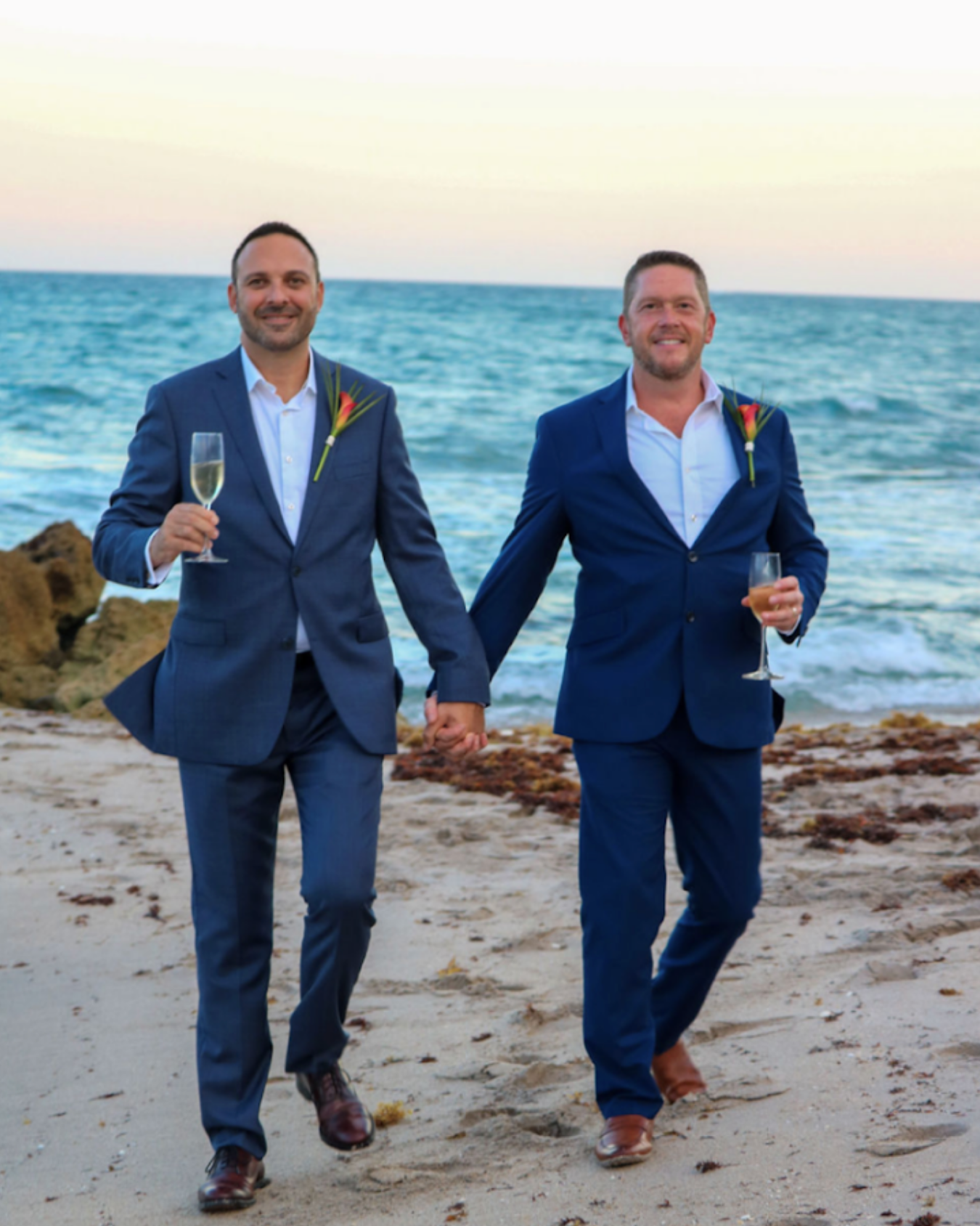 Two men in blue suits holding hands on a beach at sunset, each holding a glass of champagne, wearing boutonnières, celebrating a wedding or special occasion.