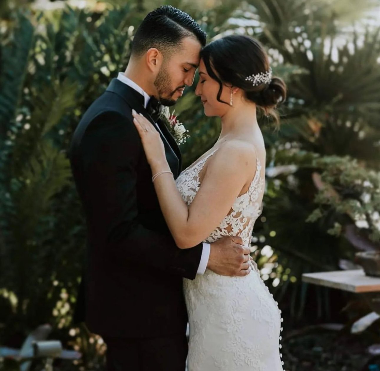 A bride and groom in a tender moment, touching foreheads, outdoors with lush greenery in the background.