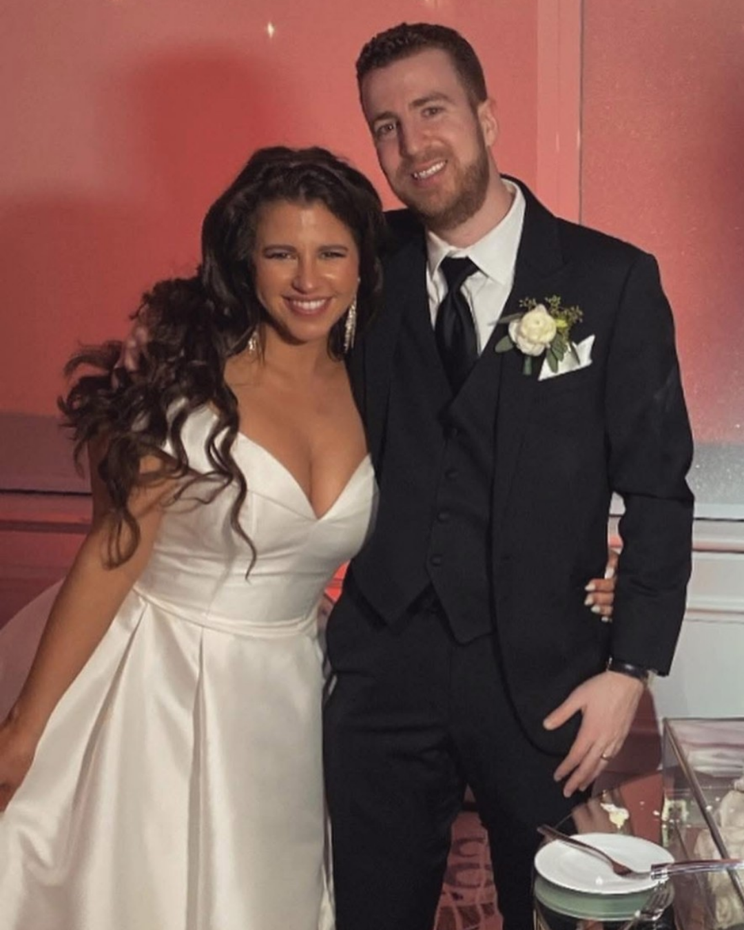 A woman in a white wedding dress and a man in a black tuxedo with a boutonniere posing together at a wedding reception.