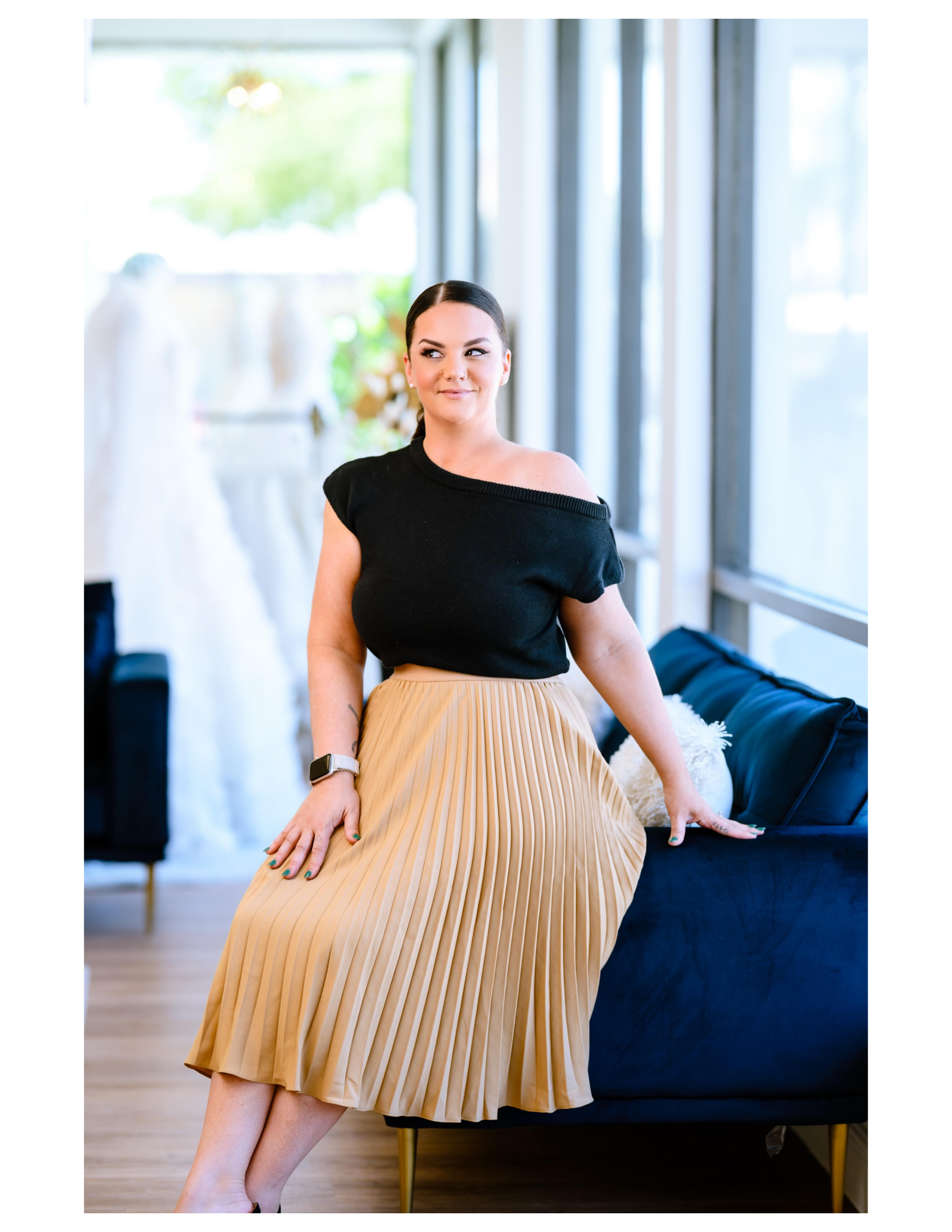 A woman sitting on a dark blue sofa in a bright room with large windows, wearing a black off-shoulder top and a beige pleated skirt, with a smartwatch on her left wrist.