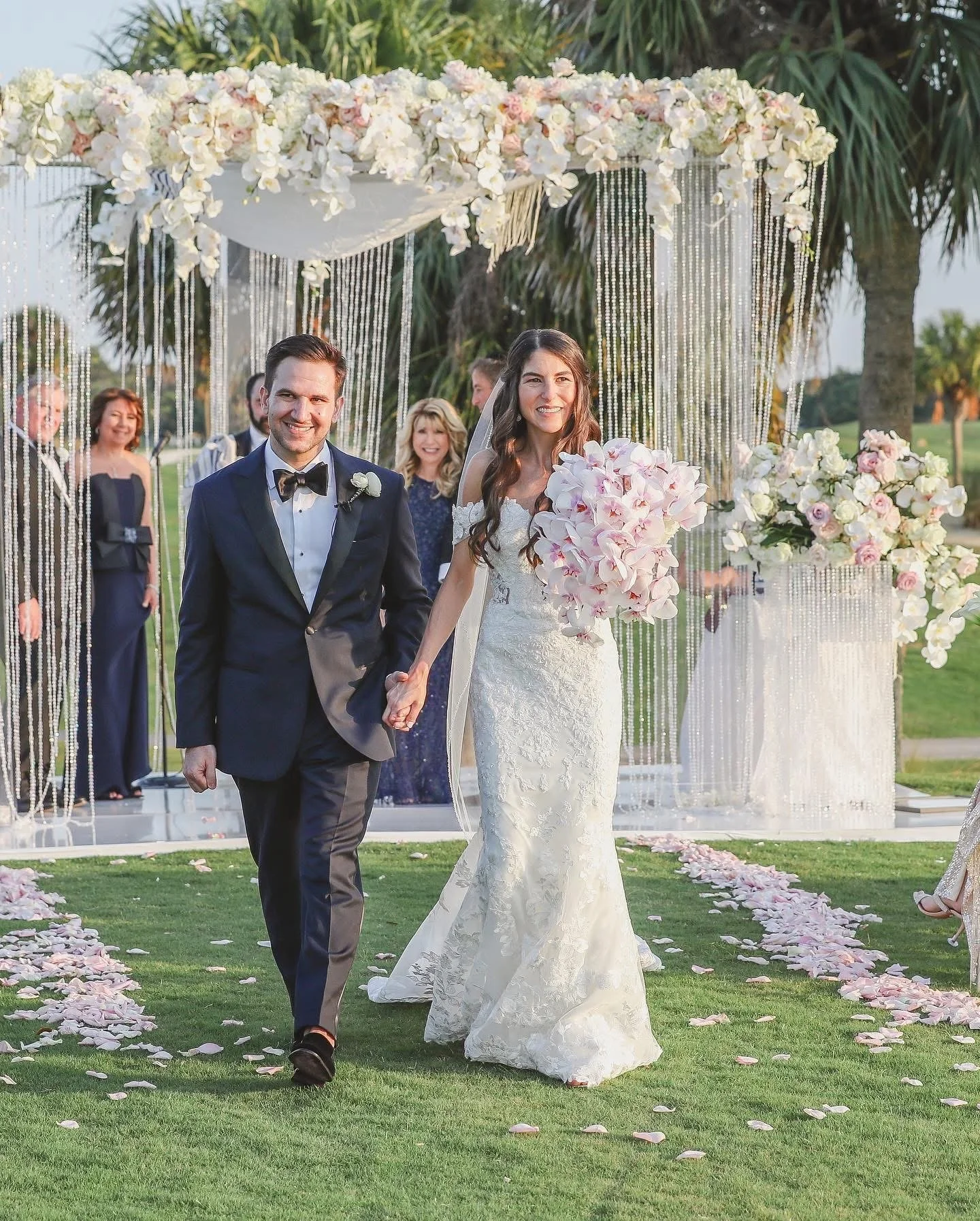 A newlywed couple walking hand in hand at their outdoor wedding ceremony, smiling, with guests and lush greenery in the background.