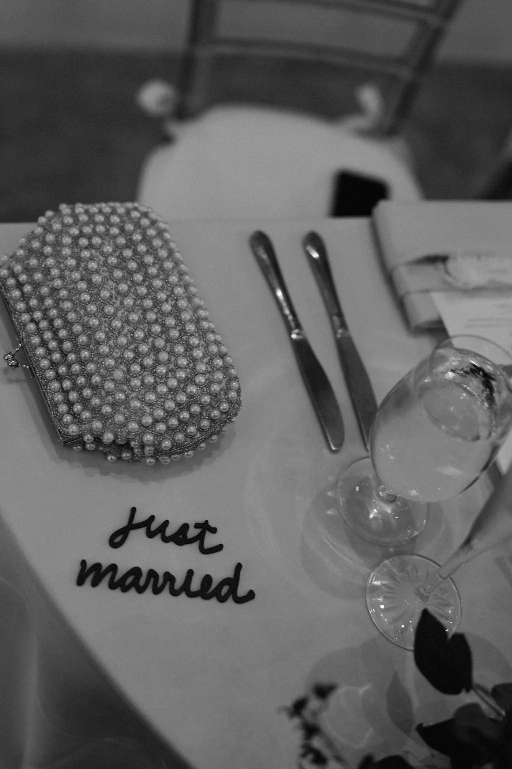 A table set for a wedding reception with a heart-shaped pearl embellished clutch, champagne glasses, and a napkin with a 