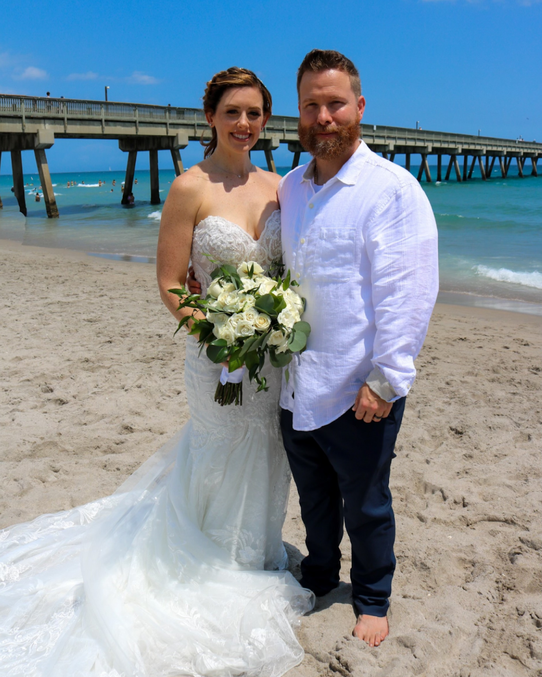 A bride and groom standing on a sandy beach with a pier in the background, a bright blue sky, and the ocean. The bride is holding a bouquet of white roses, and both are smiling at the camera.