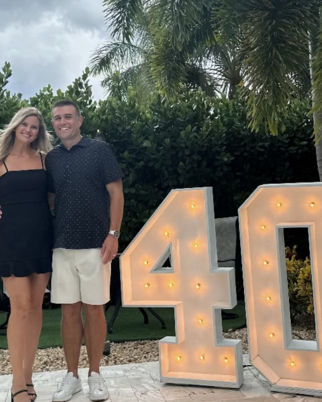 A smiling woman and man standing together outdoors near large illuminated marquee numbers '40' on a lawn with palm trees and bushes in the background.